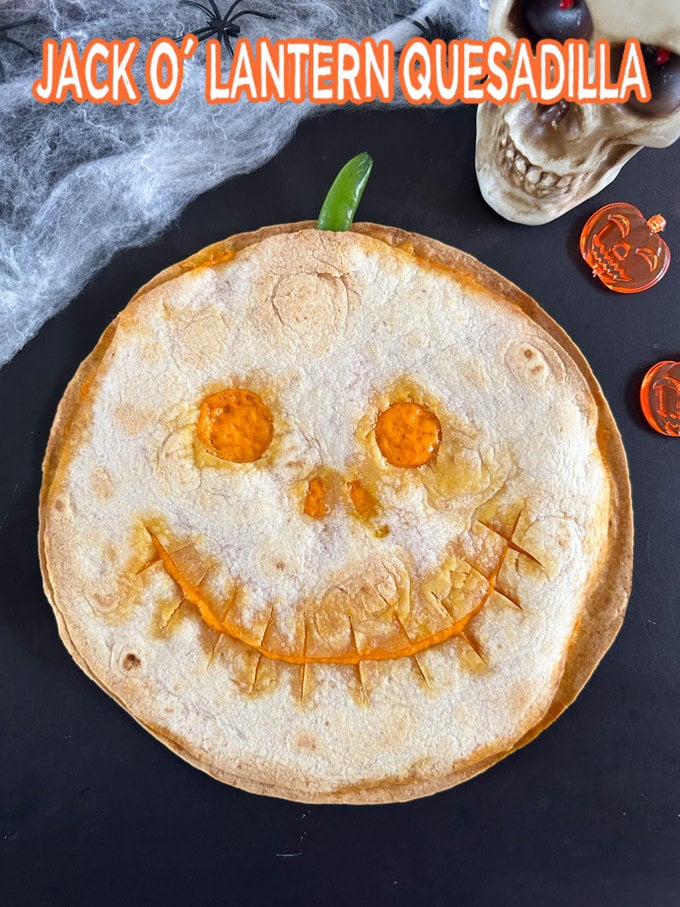 Jack O'Lantern Quesadilla on a black background with Halloween decorations in the background.