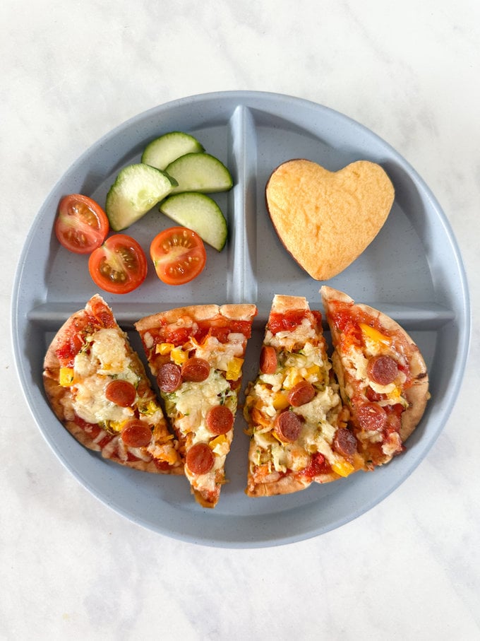 Pitta pizza bread served on a round compartment plate with sliced cucumber and cherry tomatoes, along with a small heart shaped sponge cake. 