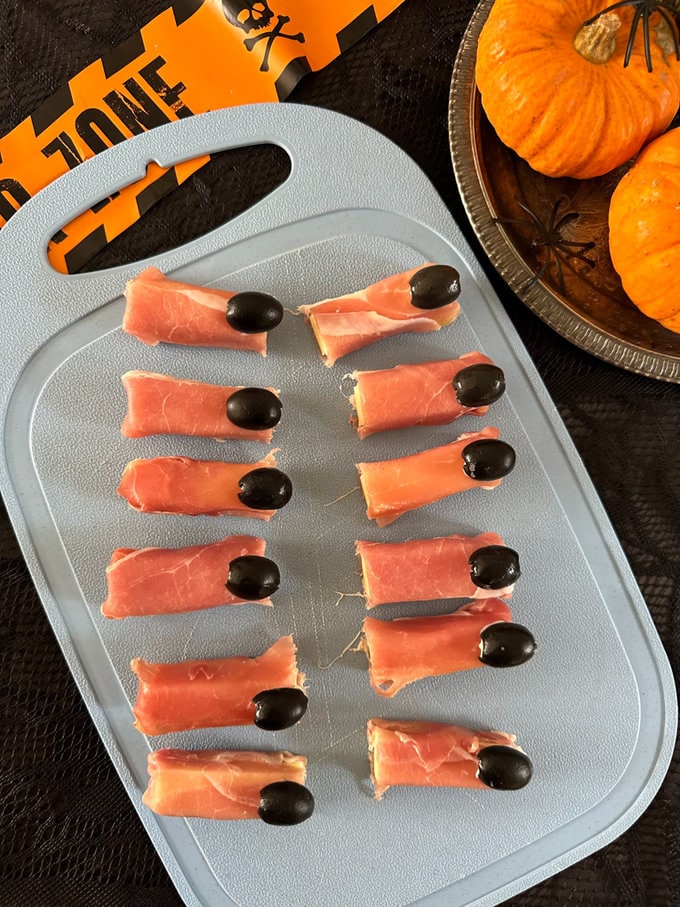Witches fingers laid out on a chopping board, surrounded by pumpkins and other Halloween decor.