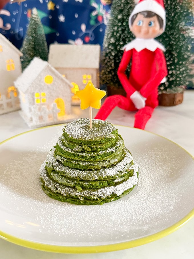 Elf on the shelf sitting next to a nice big pile of elf Christmas tree green pancakes topped with a star.