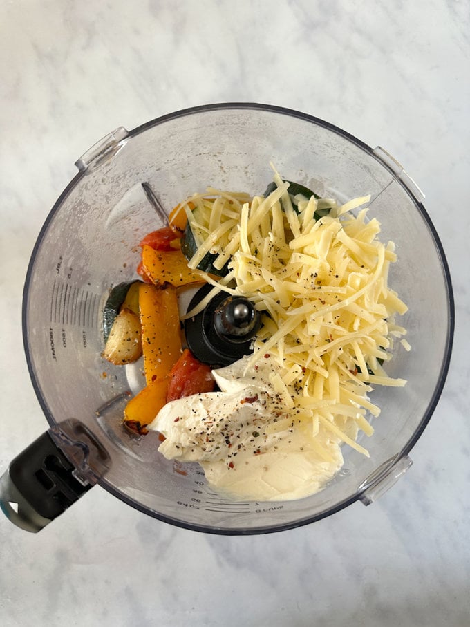 Roasted veggies being added into a food blender with grated and cream cheese.