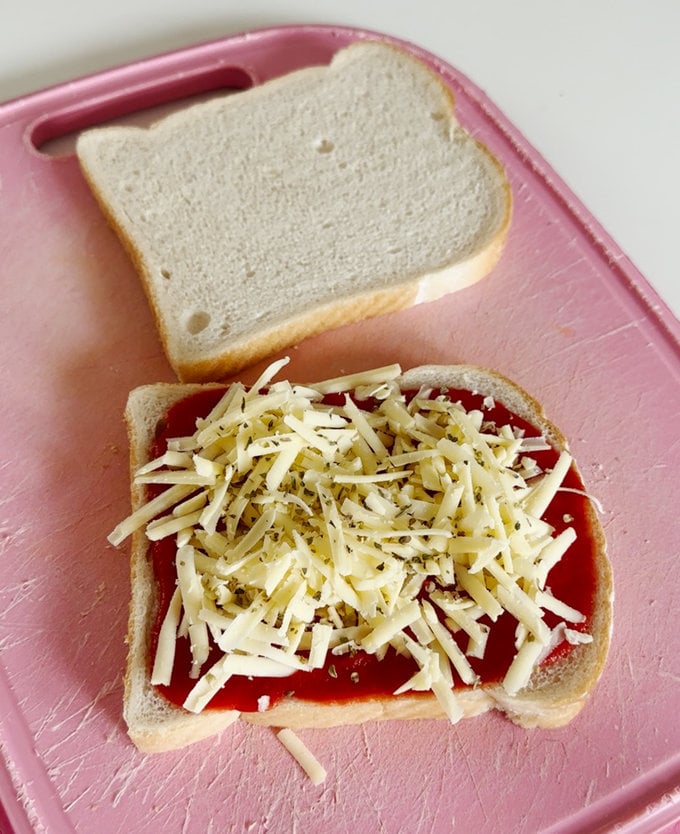 Two slices of white bread on a pink chopping board, one slice is topped with passata,grated cheddar cheese and a sprinkle of oregano.