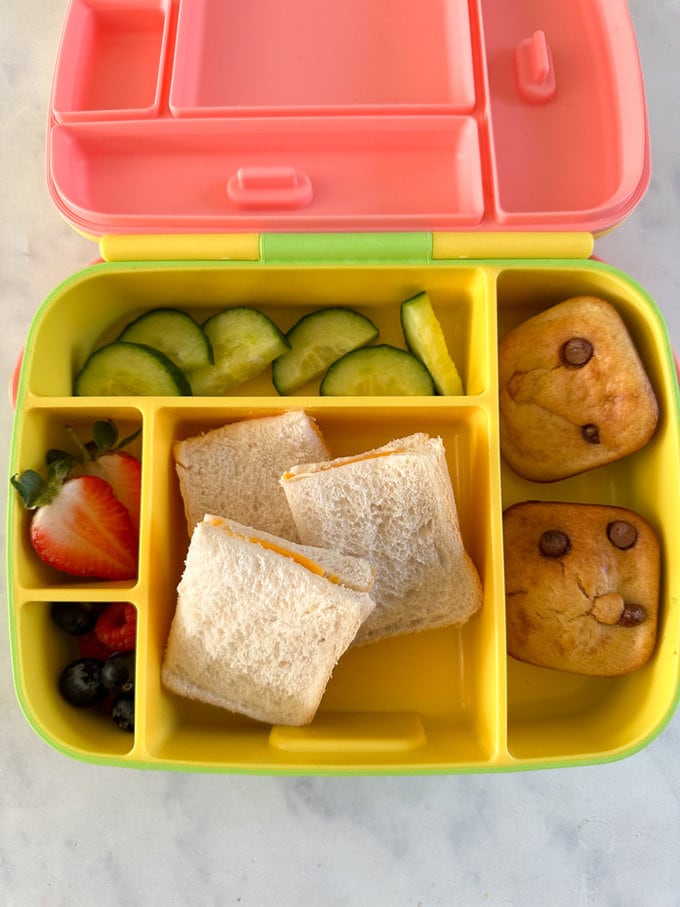 Mini Chocolate Chip Banana Bread served in a multi section bright yellow lunchbox along with three squares of sandwiches, cucumber and fresh berries.