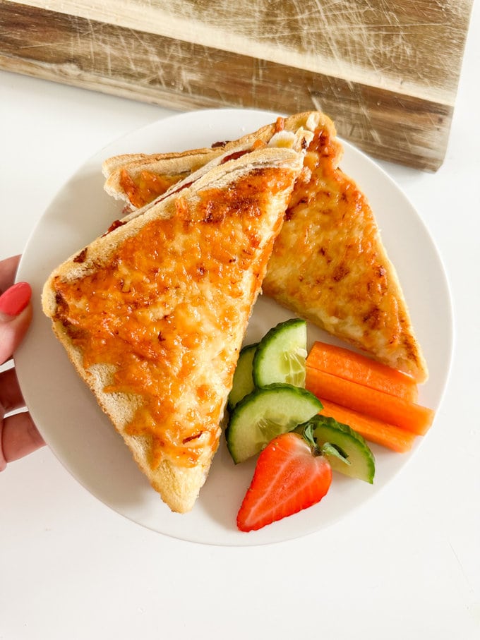Airfryer Ham & Cheese Toastie served on a white round plate, and garnished with carrot sticks,cucumber slices and half a strawberry.