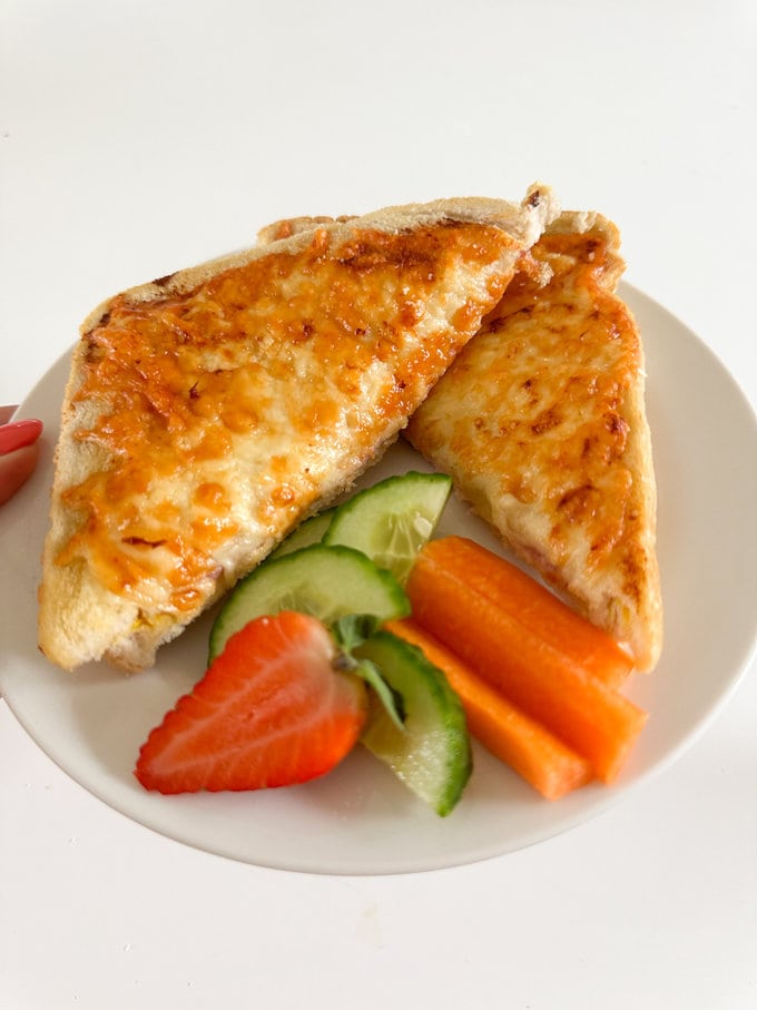 Airfryer Ham & Cheese Toastie cut into two triangles and served on a white plate with cucumber, carrot and strawberry.