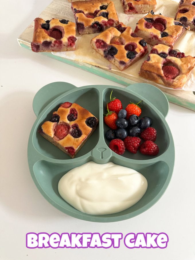 a Square of breakfast cake served in a green divided plate with a portion of yogurt and fresh summer berries.