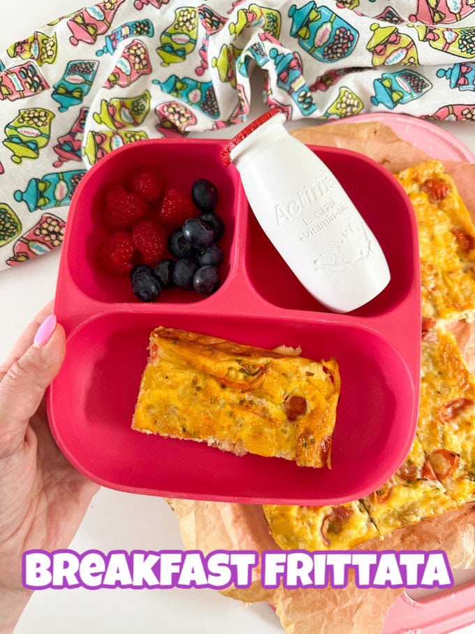 A rectangle a breakfast frittata served in a pink three compartment plate with a small bottle of Actimel and a portion of raspberries and blueberries.