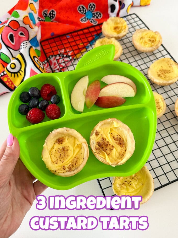 Two small custard tarts in a green apple shaped divided plate for kids with slices of apple and fresh raspberries and blueberries.