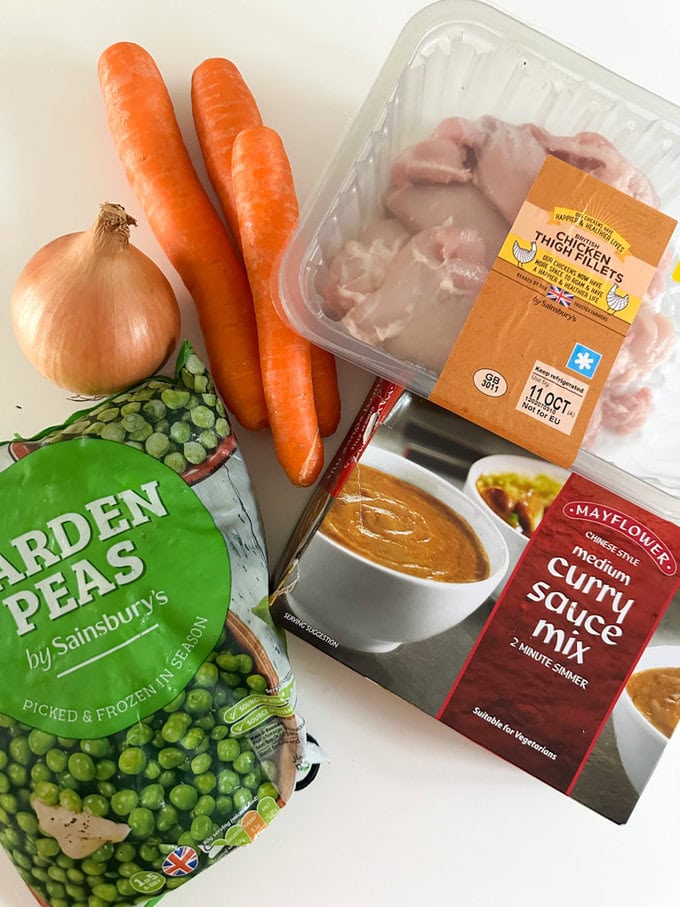 Packet of chicken thigh fillets arranged on the worktop along with Carrots, frozen peas, onion and a box of curry sauce.