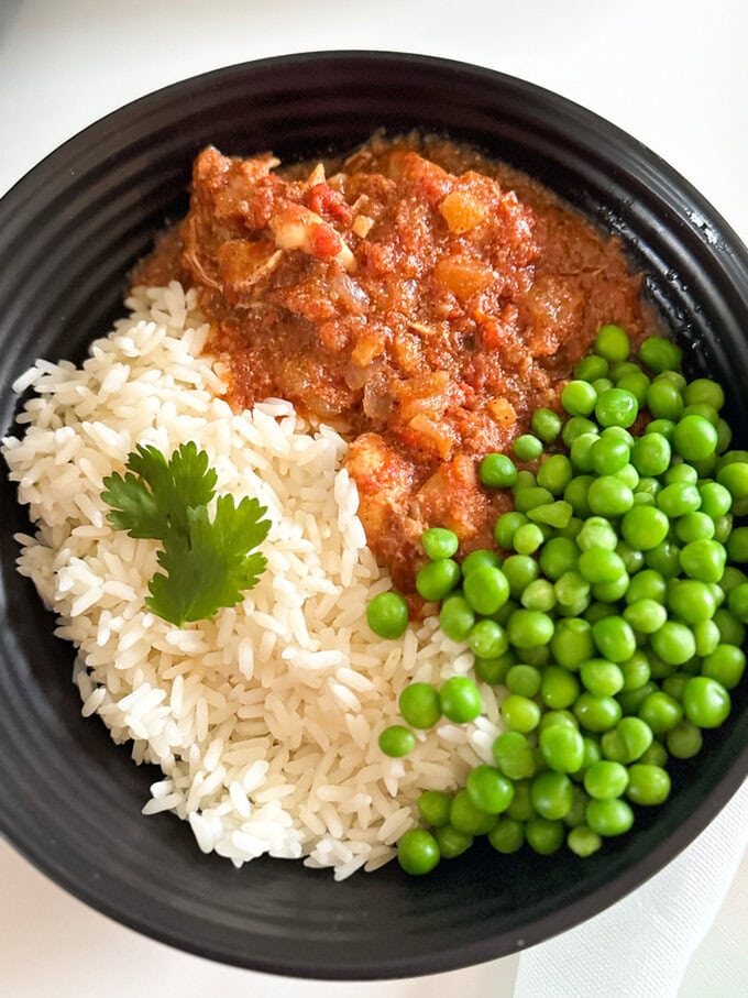 Slow Cooker Butter Chicken served in a round black dinner bowl with white rice and green peas.