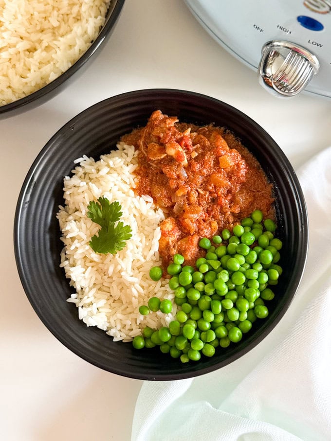 Slow Cooker Butter Chicken served in a round black dinner bowl with white rice and green peas.