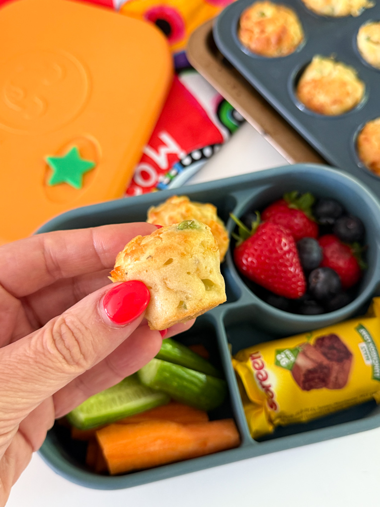 Close up of a Mini Cheese & Pea Lunchbox Muffin. Served in a bento style kids lunchbox along with fresh berries and chopped carrots and cucumber.