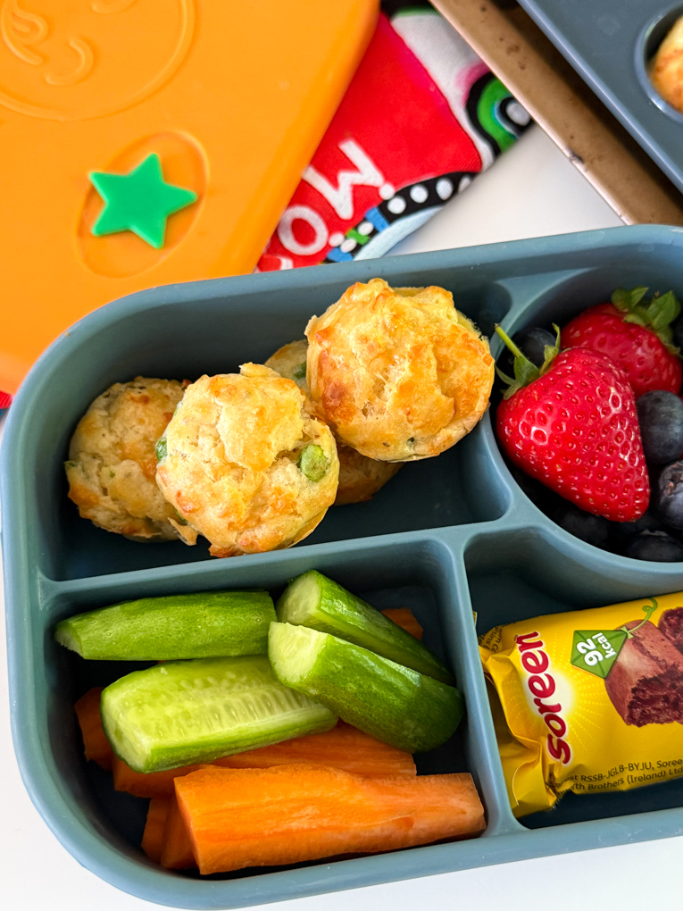 Four Mini Cheese & Pea Lunchbox Muffins served in a bento style kids lunchbox along with fresh berries and chopped carrots and cucumber.