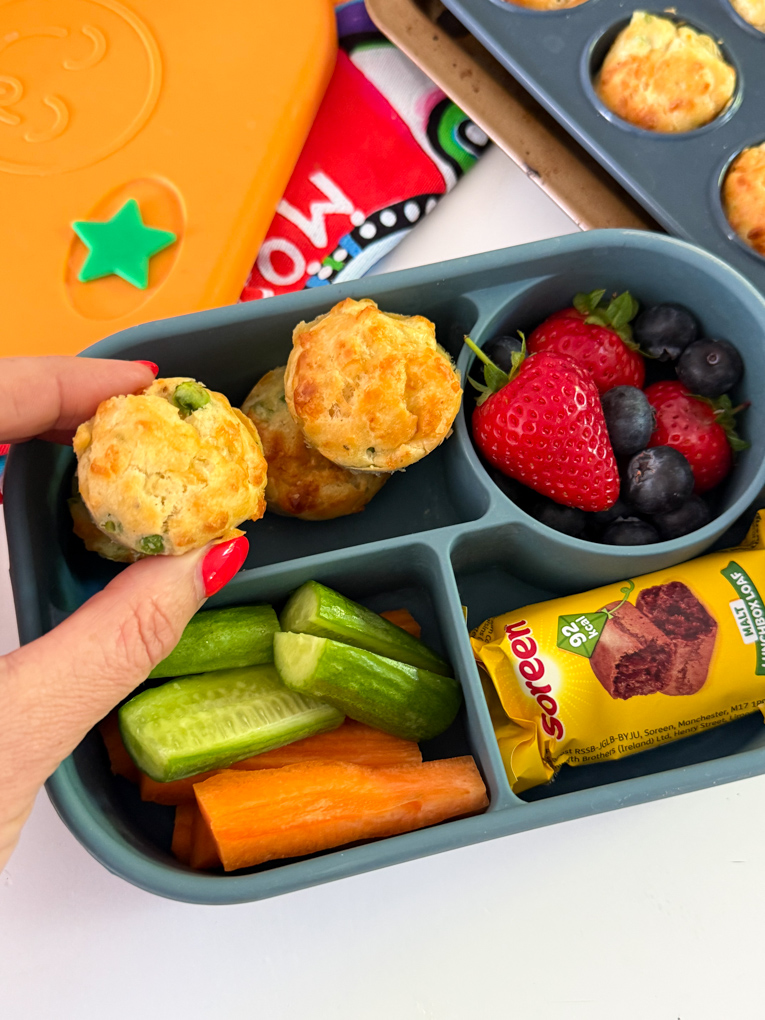 Four Mini Cheese & Pea Lunchbox Muffins served in a bento style kids lunchbox along with fresh berries and chopped carrots and cucumber.