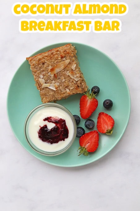 A square coconut and almond breakfast bar, served on a turquoise side plate with a small ramekin of greek yogurt and jam with chopped strawberries and blueberries on the side.