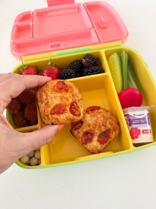 Two cheese & tomato loaves served in a kids yellow lunchbox along with chopped veggies,berries and a pot of yogurt. 