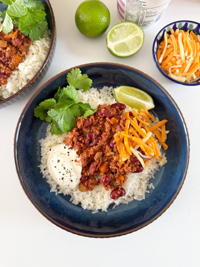 The easiest Chilli Con Carne recipe you will ever make. Just add all the ingredients to the slow cooker and leave it to cook while you get on with your day!Served with white rice,grated cheese. And garnished with a slice of lime and a sprig of coriander. In a round dark blue bowl.