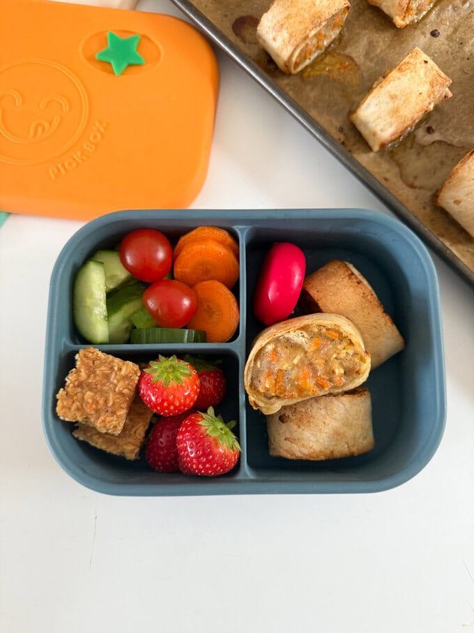 Served in a child's blue lunch box alongside fresh chopped fruit & veggies.