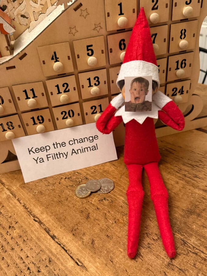 Elf sat against a wooden advent calendar, with a cut out of Kevin from home alone stuck over the elf's face, with a note beside saying keep the change ya flithy animal, with a few coins underneath.
