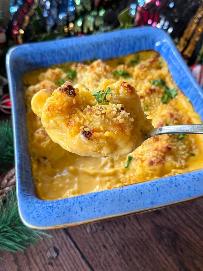 Make Ahead Cauliflower Cheese served in a bright blue oven dish, one piece being scooped up with a large spoon.