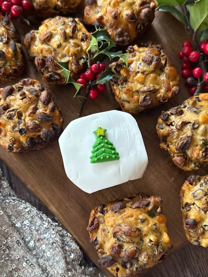 Mini Christmas cakes arranged on a wooden serving board decorated with holly and berries. One if cakes is decorated with white fondant icing with a small Christmas tree in the middle.
