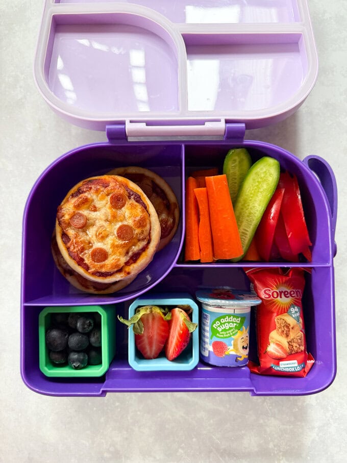Three mini pizzas in a kids divided purple lunchbox, served with fresh chopped veggies & berries and some other tasty snacks.