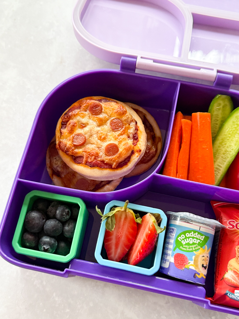Three mini pizzas in a kids divided purple lunchbox, served with fresh chopped veggies & berries and some other tasty snacks.