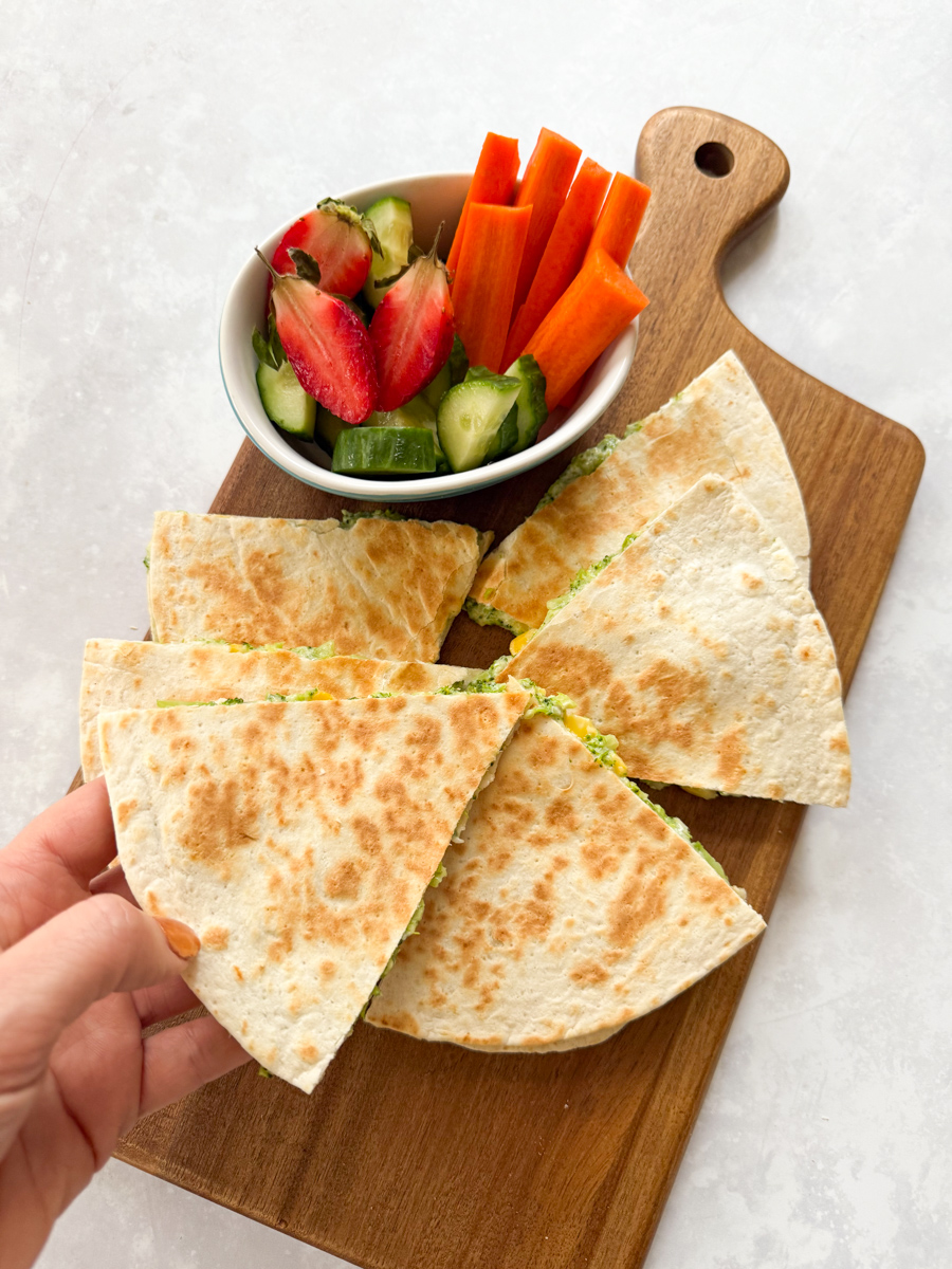 Broccoli quesadillas arranged on a wooden chopping board, with a small bowl of fresh chopped fruit & veggies.