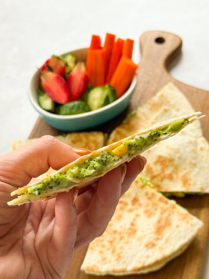 Broccoli quesadillas arranged on a wooden chopping board, with a small bowl of fresh chopped fruit & veggies.