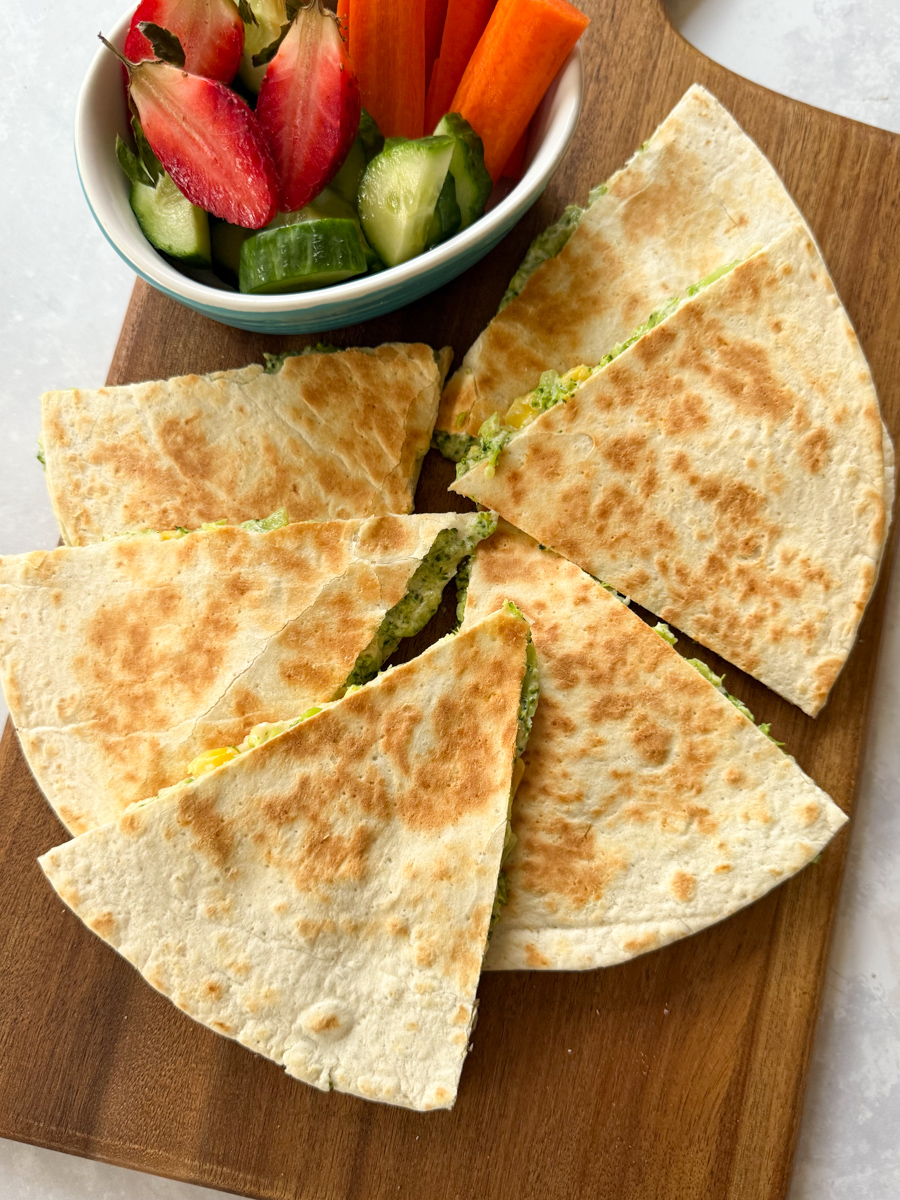 Broccoli quesadillas arranged on a wooden chopping board, with a small bowl of fresh chopped fruit & veggies.