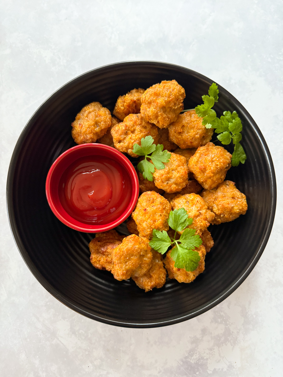 Chicken nuggets with veggies served in a large round,black bowl with a separate small red bowl filled with ketchup ready for dipping.