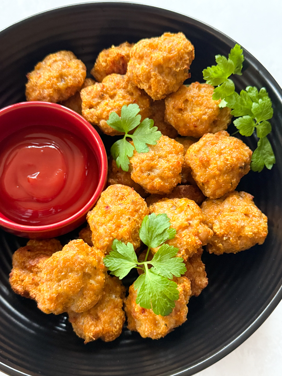 Chicken nuggets with veggies served in a large round,black bowl with a separate small red bowl filled with ketchup ready for dipping.