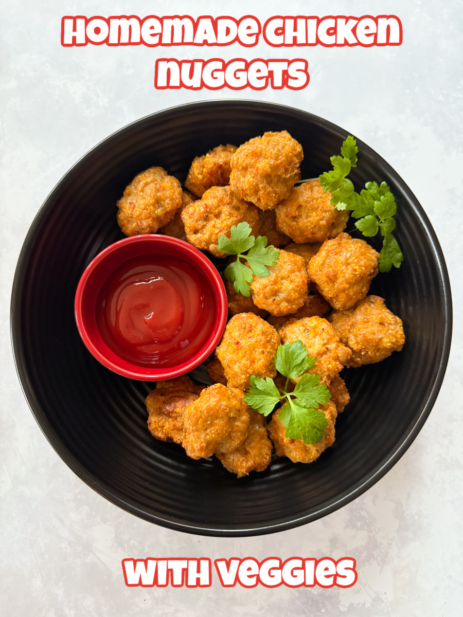 Chicken nuggets with veggies served in a large round,black bowl with a separate small red bowl filled with ketchup ready for dipping.