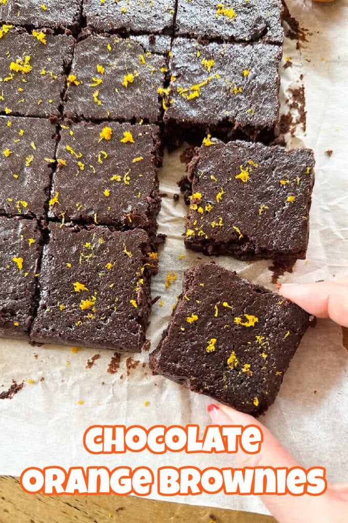 A top-down view of chocolate orange brownies that have been cut into squares on a sheet or parchment paper garnished with orange zest and a female handing taking a brownie portion.