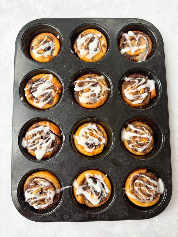 Petits pains à la cannelle disposés dans un moule à muffins, dorés et recouverts de glaçage blanc.