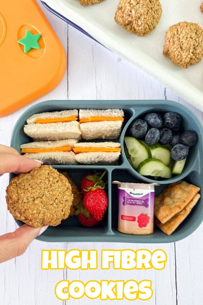 A top-down view of a blue silicone bento box containing a cheese sandwich, blueberries, chopped cucumber, strawberries, a small yoghurt pot, dry crackers and a female hand holding a high fibre cookie above the lunchbox.
