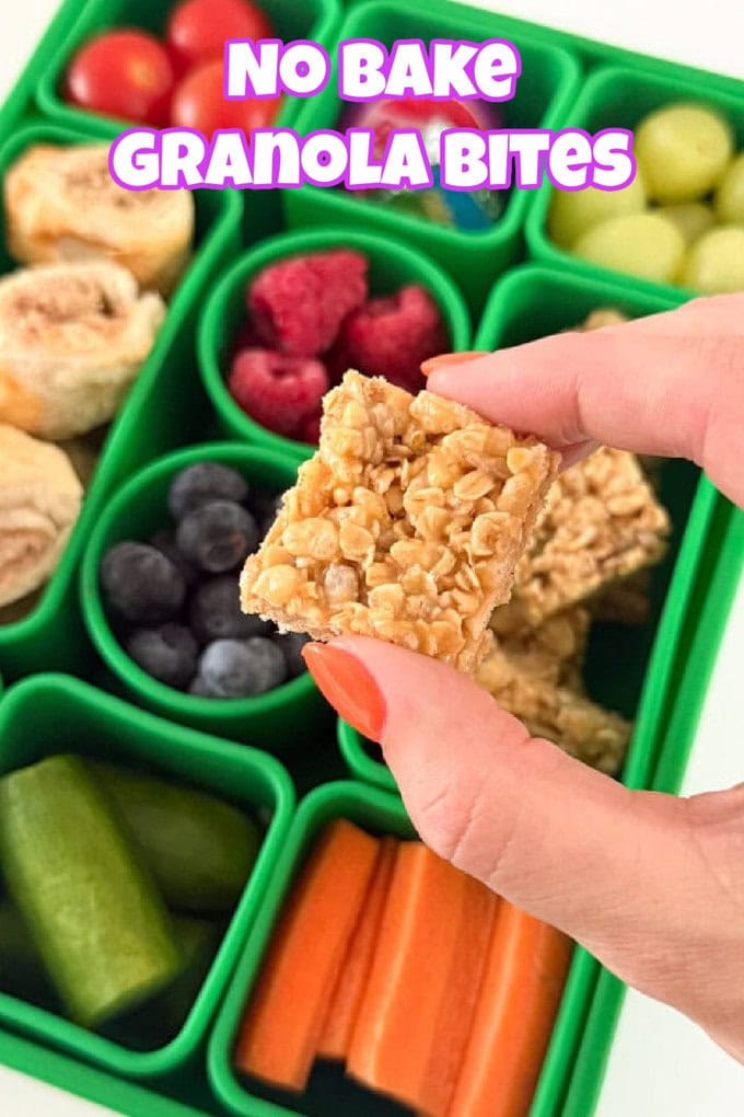 A top-down shot of a female hand holding a mini no bake granola bite above a green silicone snackle box from Pick Plates filled with healthy fruit and veg snacks.