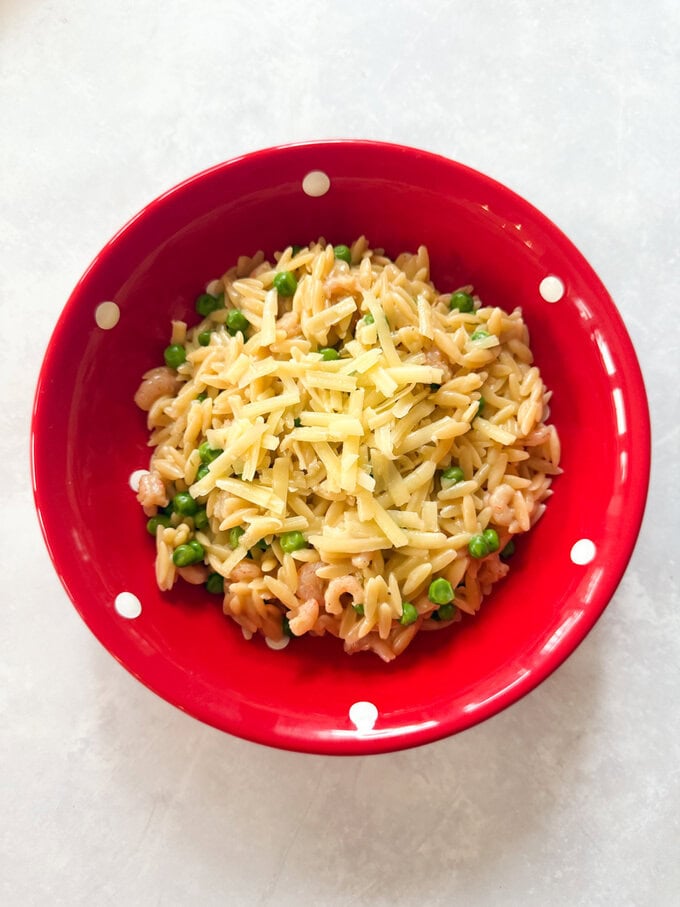 Un morceau d'orzo aux crevettes servi dans un bol rouge vif à pois blancs, avec une cuillère vert vif à côté du bol, un orzo garni de fromage râpé.