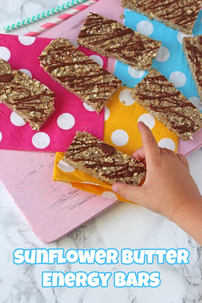A top-down view of a child's hand taking a chocolate drizzled energy bar from a pink wooden chopping board that has brightly coloured napkins on it and more slices of the energy bars.