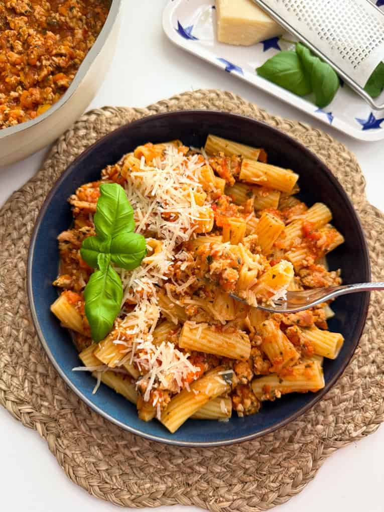 Chicken Mince Bolognese served in a dark blue large round pasta bowl, garnished with grated cheese and a sprig of basil.