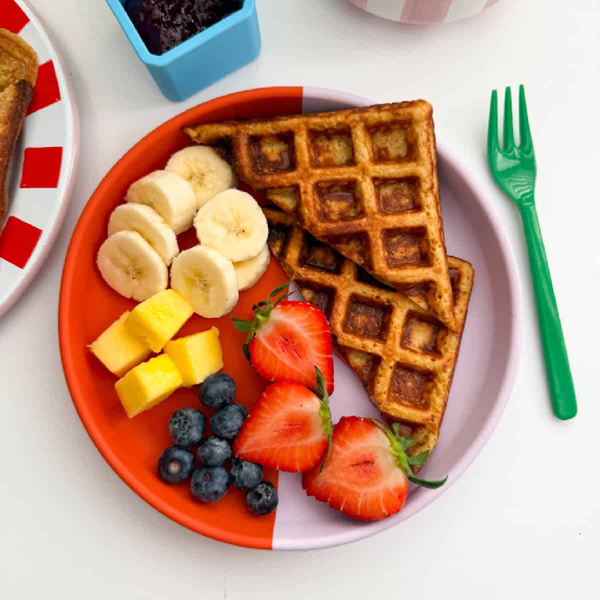 Porridge oat waffle served on a bright red and pink small round plate with a bright green folk, and served with fresh berries and sliced banana.