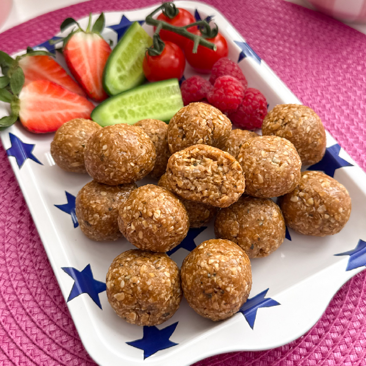 A pile of Biscoff Oat Energy Bites served on a white tray with a blue star pattern, along with fresh,chopped berries and cucumber.