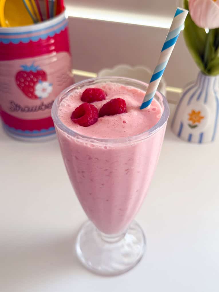 Frozen Raspberry Shake in a tall milkshake glass topped with fresh raspberries and a blue and white striped straw.