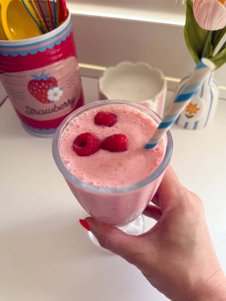 Frozen Raspberry Shake in a tall milkshake glass topped with fresh raspberries and a blue and white striped straw.
