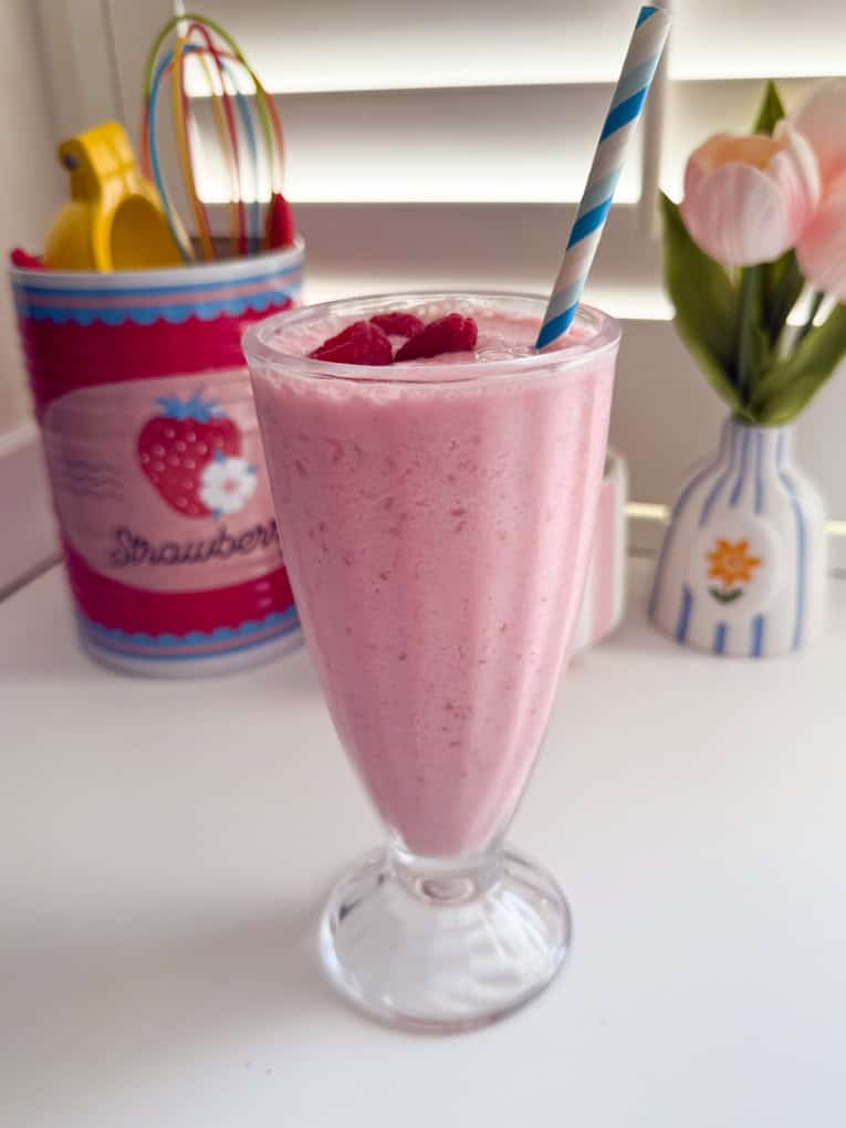 Frozen Raspberry Shake in a tall milkshake glass topped with fresh raspberries and a blue and white striped straw.