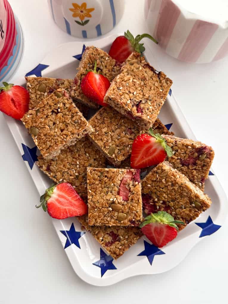 Strawberry oat bars cut into bite size squares and served on a rectangular shaped dish with a bright blue star pattern all over, garnished with fresh strawberries.